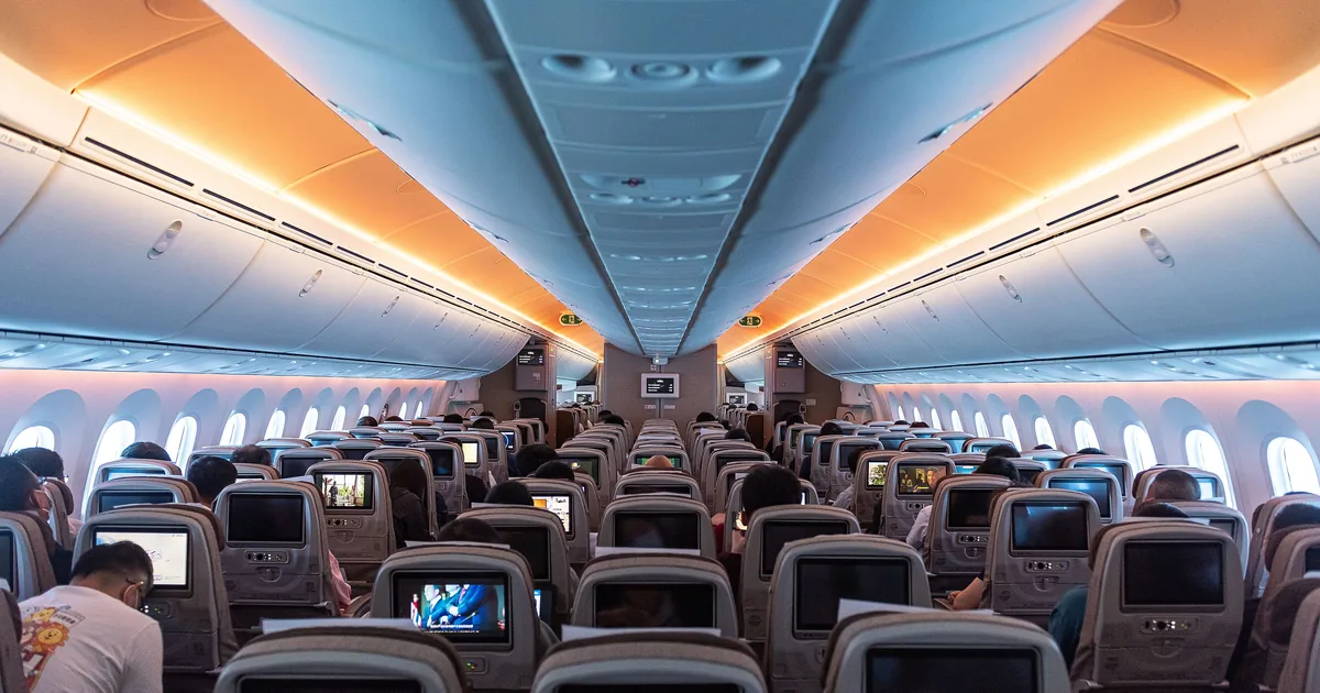 Cabina de clase turista de un avión en vuelo, con los maleteros cerrados a ambos lados y pasajeros en sus asientos.